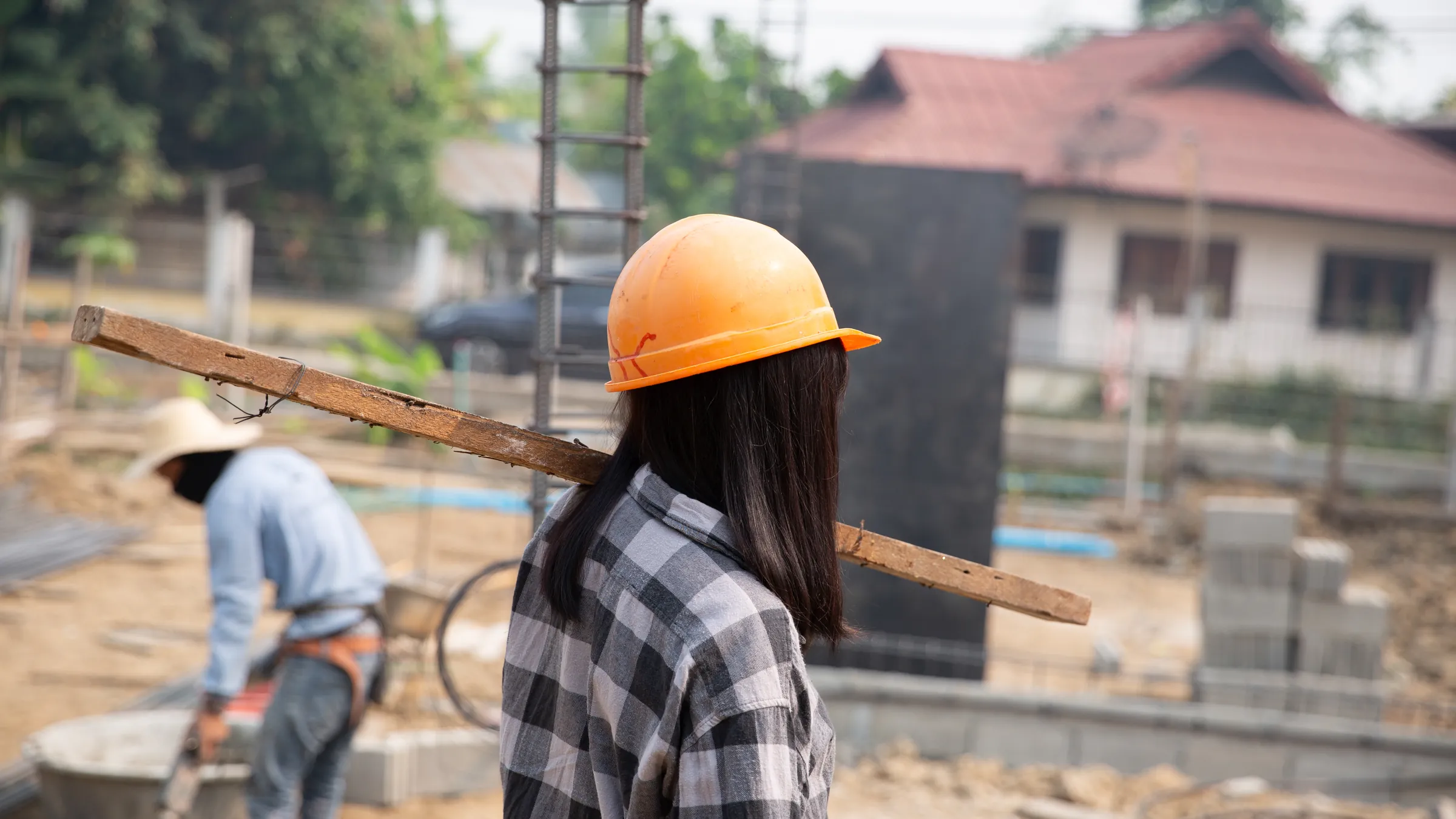 Builders in hard hats carrying timber across a house construction site with block walls rising