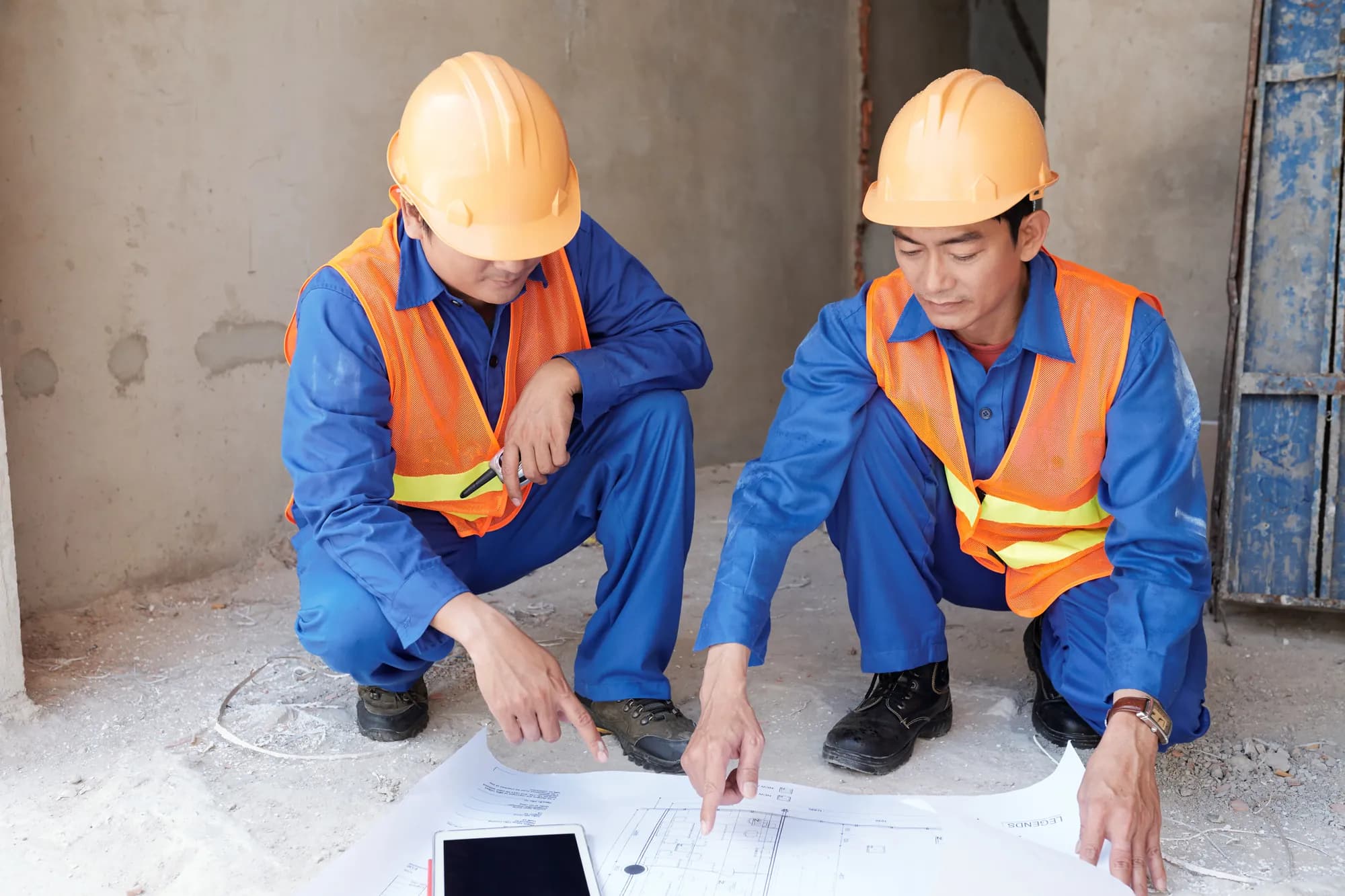 Two engineers in hard hats and safety vests kneeling over construction drawings on a site floor