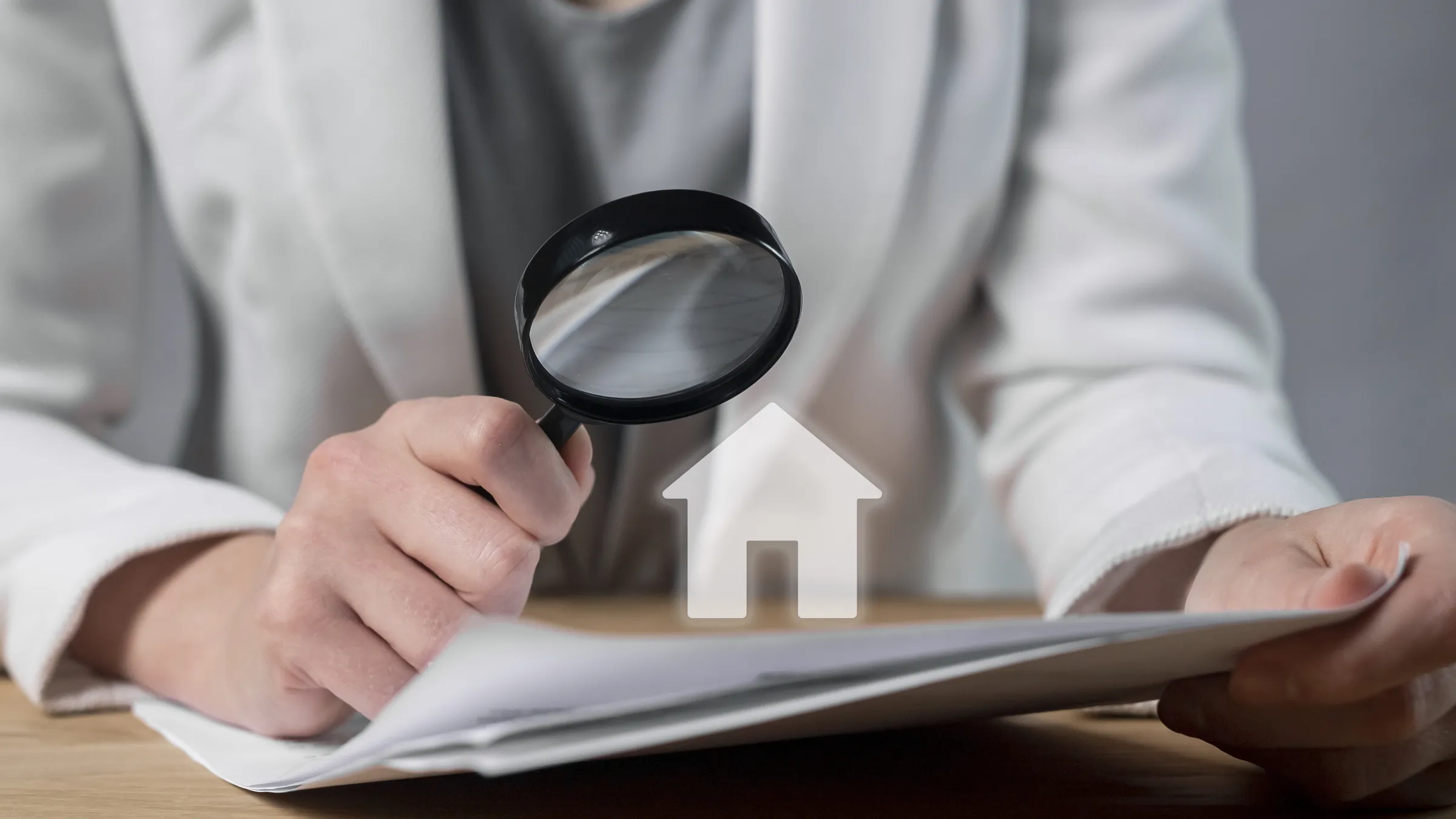 Hands holding a magnifying glass over a property purchase agreement, next to a small model house